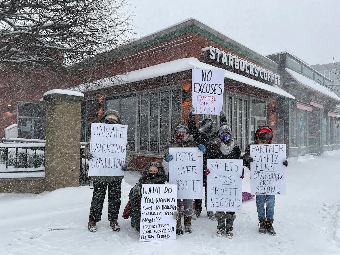 Starbucks workers picketing outside a store in heavy snow, holding hand-lettered signs reading "Unsafe working conditions", "People over profit", "Safety first, profit second" and "No excuses — employee safety first".