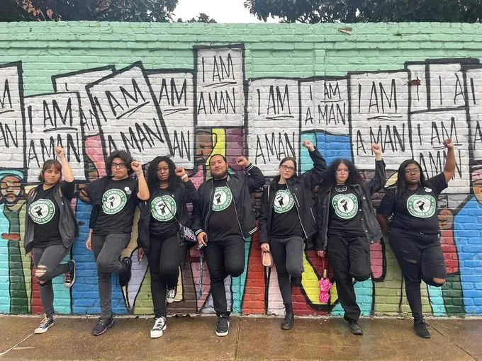 The Memphis 7 — seven Starbucks Workers United members in union shirts, fists raised, standing in front of a mural of the 1968 Memphis sanitation strike's "I AM A MAN" placards.