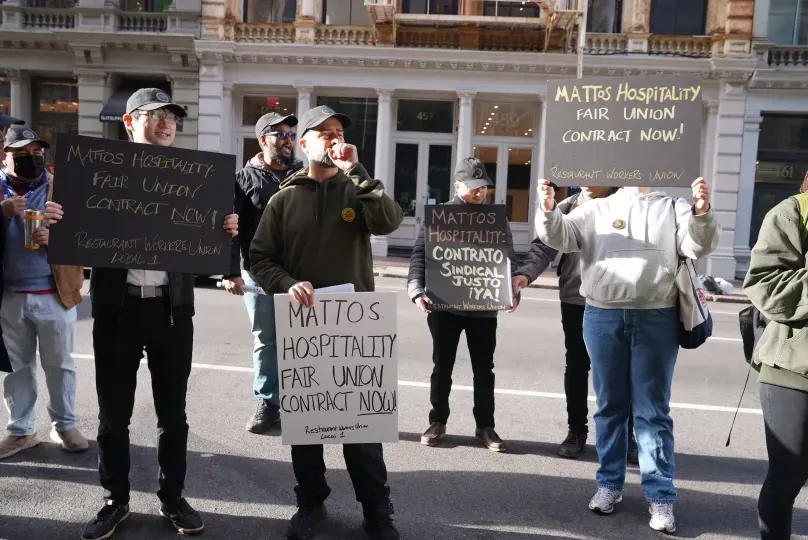Una fila de trabajadores de restaurantes en una acera de Manhattan con carteles negros hechos a mano. Uno dice "Mattos Hospitality: Fair Union Contract Now!" y otro, en español, "Mattos Hospitality: Contrato Sindical Justo ¡Ya!"