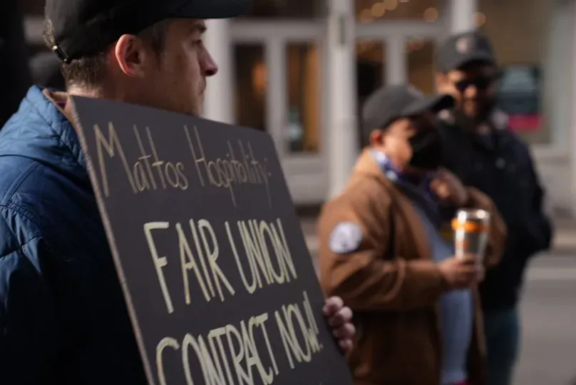 Close-up of a worker holding a hand-painted sign reading "Mattos Hospitality: Fair Union Contract Now!", with other picketers out of focus behind.