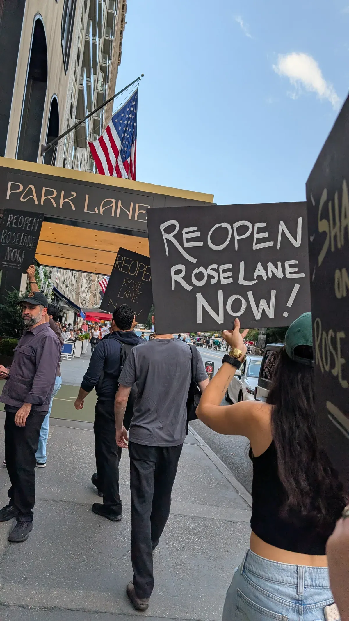 A worker holds a hand-painted sign reading "Reopen Rose Lane Now!" above her head on the sidewalk outside the Park Lane Hotel.