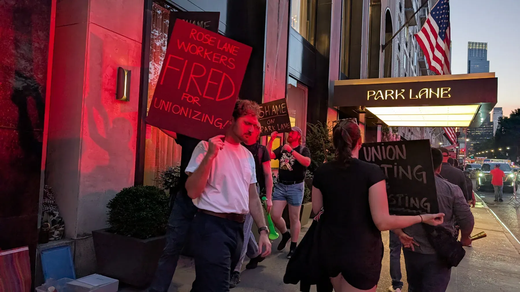 Manifestantes afuera del Park Lane Hotel al anochecer, con la fachada iluminada de rojo. Un cartel dice "Rose Lane Workers Fired For Unionizing!"