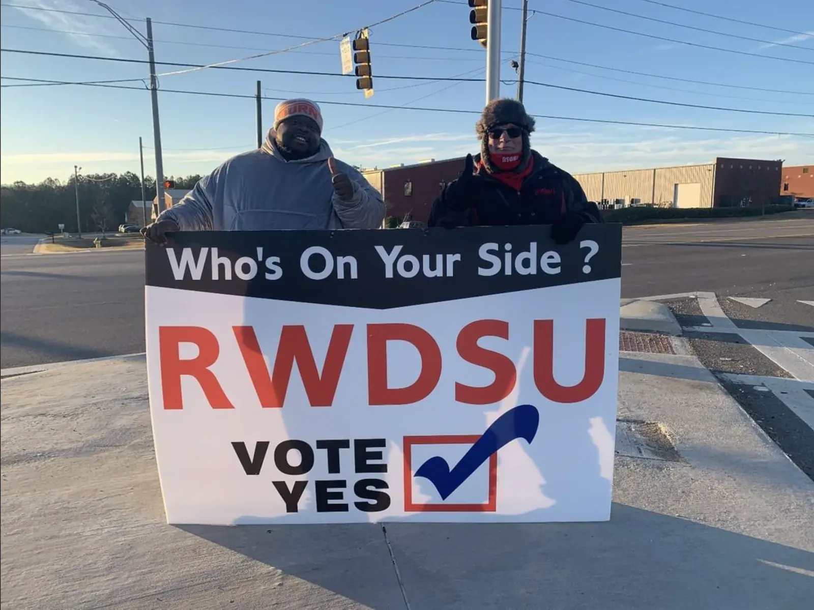 Two Amazon warehouse workers at a roadside intersection holding a large RWDSU campaign sign reading "Who's On Your Side? RWDSU — Vote Yes".